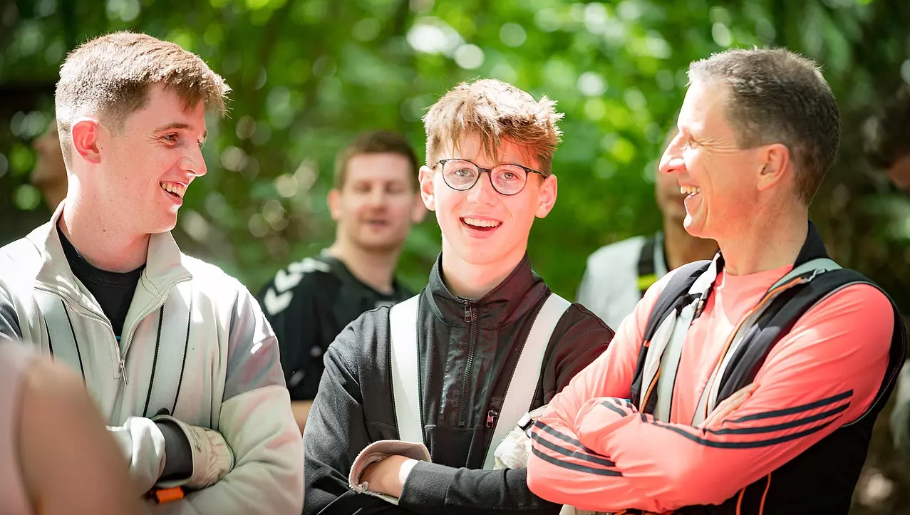 Drei Männer stehen im Freien, lachen und unterhalten sich. Zwei von ihnen sind jünger, einer älter. Im Hintergrund sind weitere Personen sowie grüne Bäume zu sehen. Die Stimmung wirkt locker und freundlich. Drei Männer stehen im Freien, lachen und unterhalten sich. Zwei von ihnen sind jünger, einer älter. Im Hintergrund sind weitere Personen sowie grüne Bäume zu sehen. Die Stimmung wirkt locker und freundlich.